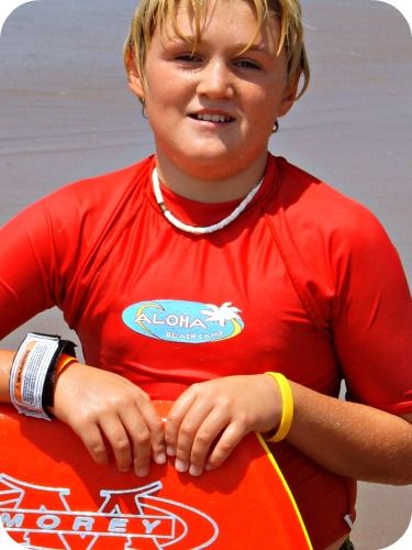 Boy standing on beach wearing red rashguard and puka shell necklace. Red boogie board in hand.