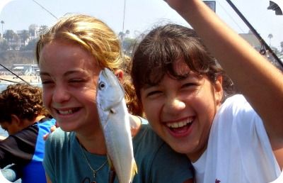Two High Action camper girls, each 13 years old, standing on the Santa Monica Pier smiling while proudling showing off the small fish they caught at Aloha Beach Camp's fishing derby activity day.