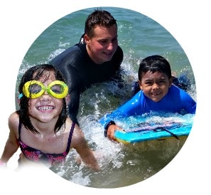 Two happy Keiki Campers, one 5 year old girl and a 6 year old boy, swim and boogie board together in the ocean as their Camp Counselor, Jeremy Polon, looks on.
