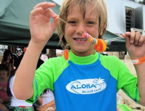 Young boy in green and blue Aloha Beach Camp rashguard proudly showing off is candy lei he made at camp.