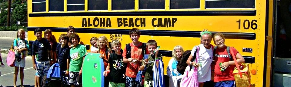 Campers pose with their beach towels, boogie boards and other camp gear in front of an Aloha Beach Camp summer camp bus.